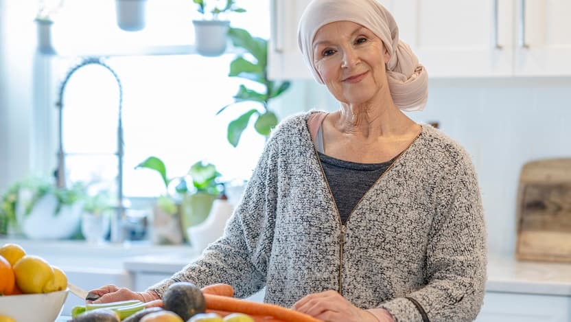 An elderly female cancer patient stands at a kitchen counter with some vegetables in front of her