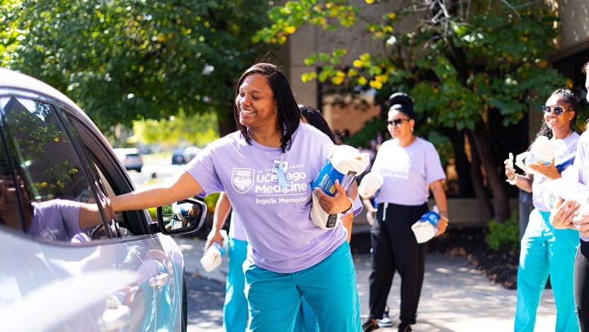 A smiling UChicago employee in a purple shirt hands baby supplies through a car window; other staff in matching purple shirts are smiling and holding more supplies.
