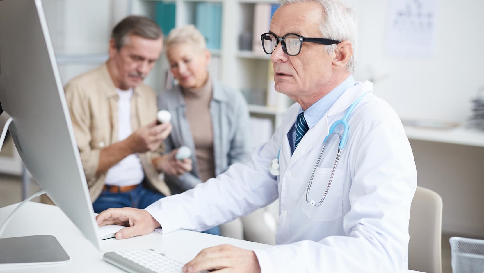 A doctor in a white coat uses a computer intently while a couple of patients examine pill bottles in the background.