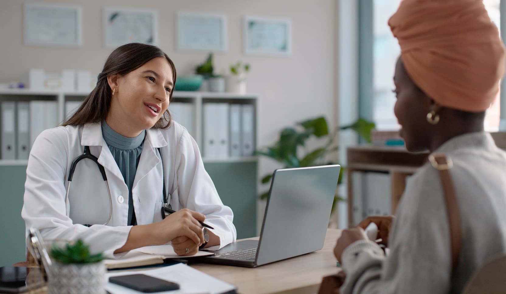 A doctor in a white coat with a stethoscope around her neck and a laptop computer in front of her sits across the desk from a patient while they converse