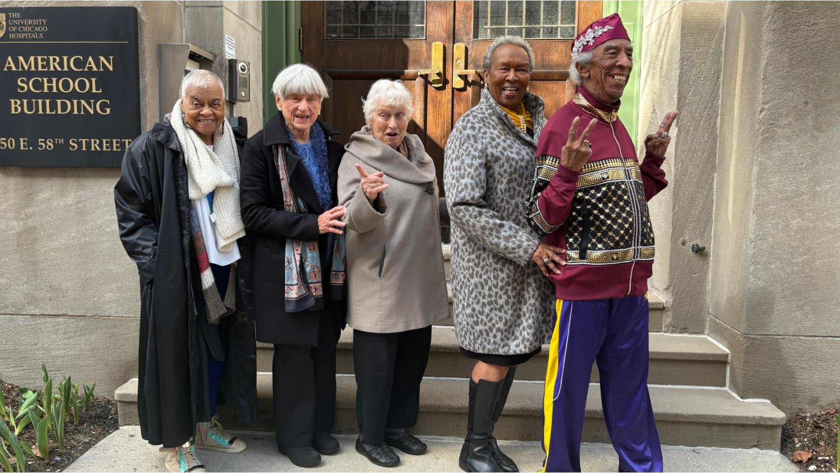 A group of five well-dressed elderly people stand smiling in a line in front of the American School Building at UChicago